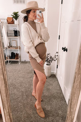 woman wearing a beige maternity dress with a white knit cardigan and holding her baby bump in front of a mirror