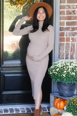 woman wearing a beige ribbed maternity dress and brown hat, holding her baby bump in front of a door decorated for fall