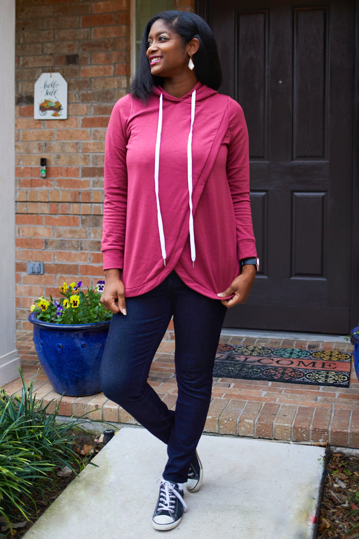 woman wearing a maroon hooded long sleeve top with white drawstrings and dark jeans, standing outside a house