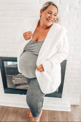 woman wearing a cozy white maternity cardigan over a grey tank top and matching leggings, showing off her baby bump