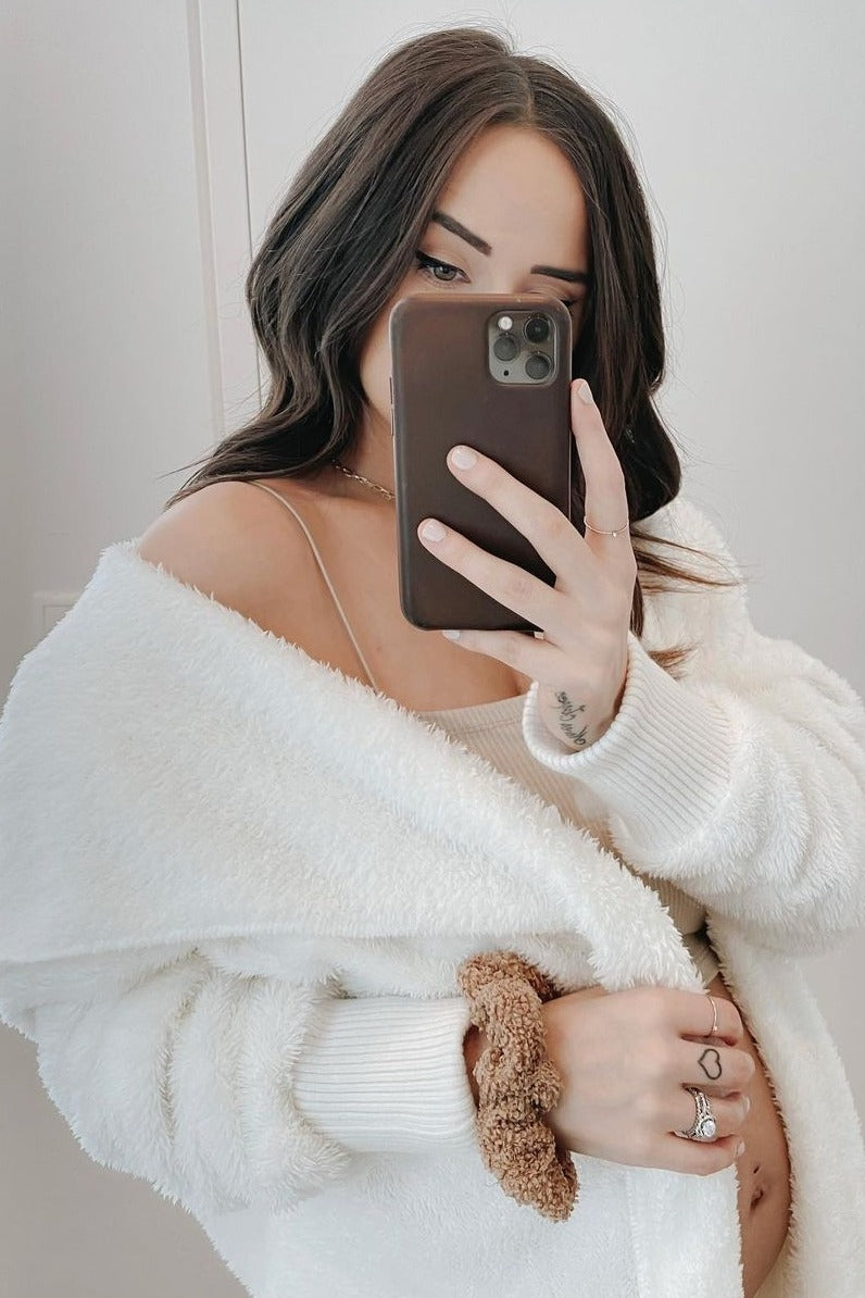 woman wearing a white cozy maternity cardigan and a light top, holding her baby bump while taking a selfie in a mirror