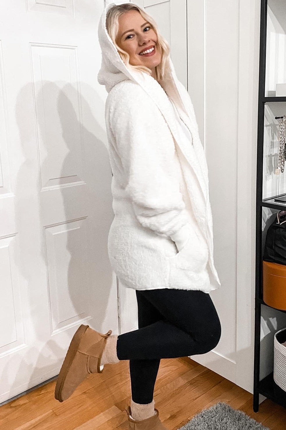 woman wearing a white fluffy hoodie coat with a hood, black leggings, and ankle boots, smiling indoors