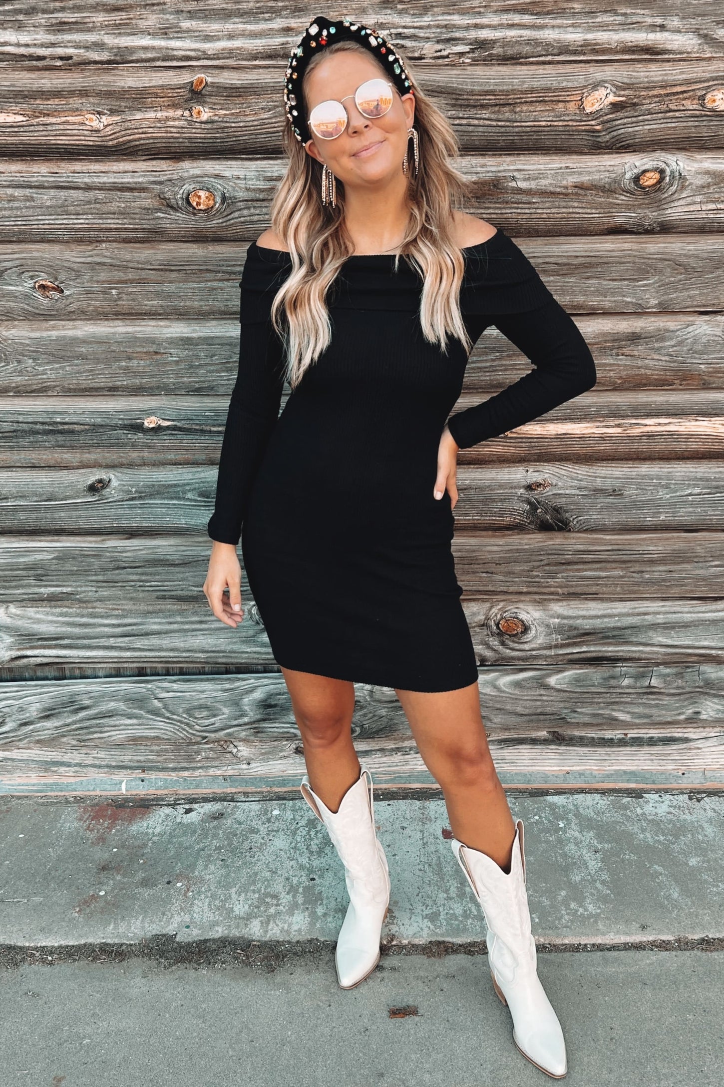 woman wearing a black off-shoulder bodycon dress, white cowboy boots, and round sunglasses, standing against a wooden backdrop