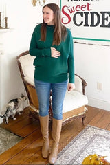 woman wearing a green maternity sweater, holding her baby bump, paired with skinny jeans and tall brown boots