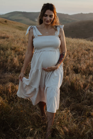 Pregnant woman in a white smocked dress walking through a golden field at sunset with hills in the background