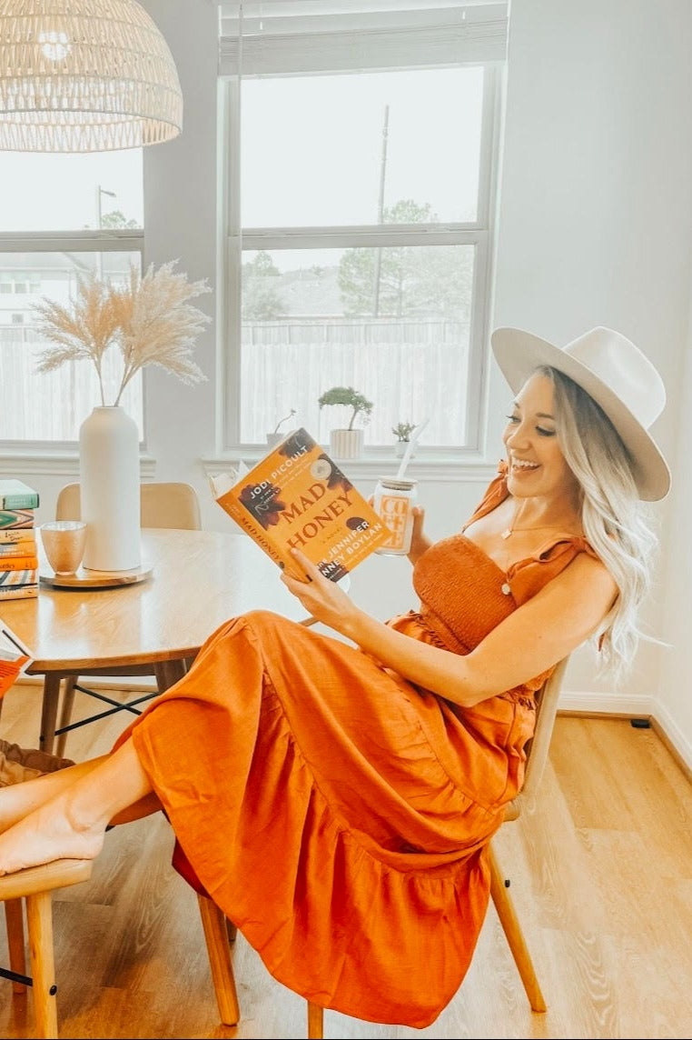 woman sitting at a table wearing an orange tiered midi dress and a wide-brimmed hat, reading a book