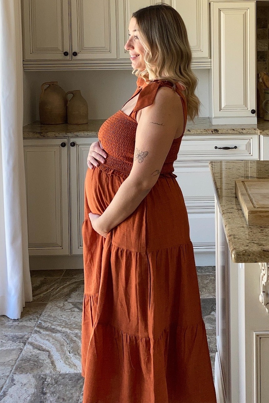 woman wearing an orange maternity maxi dress with a smocked bodice and tiered skirt, gently cradling her baby bump