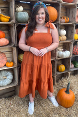 woman wearing an orange tiered sundress with tie straps and white sneakers, posing among pumpkins in a rustic setting