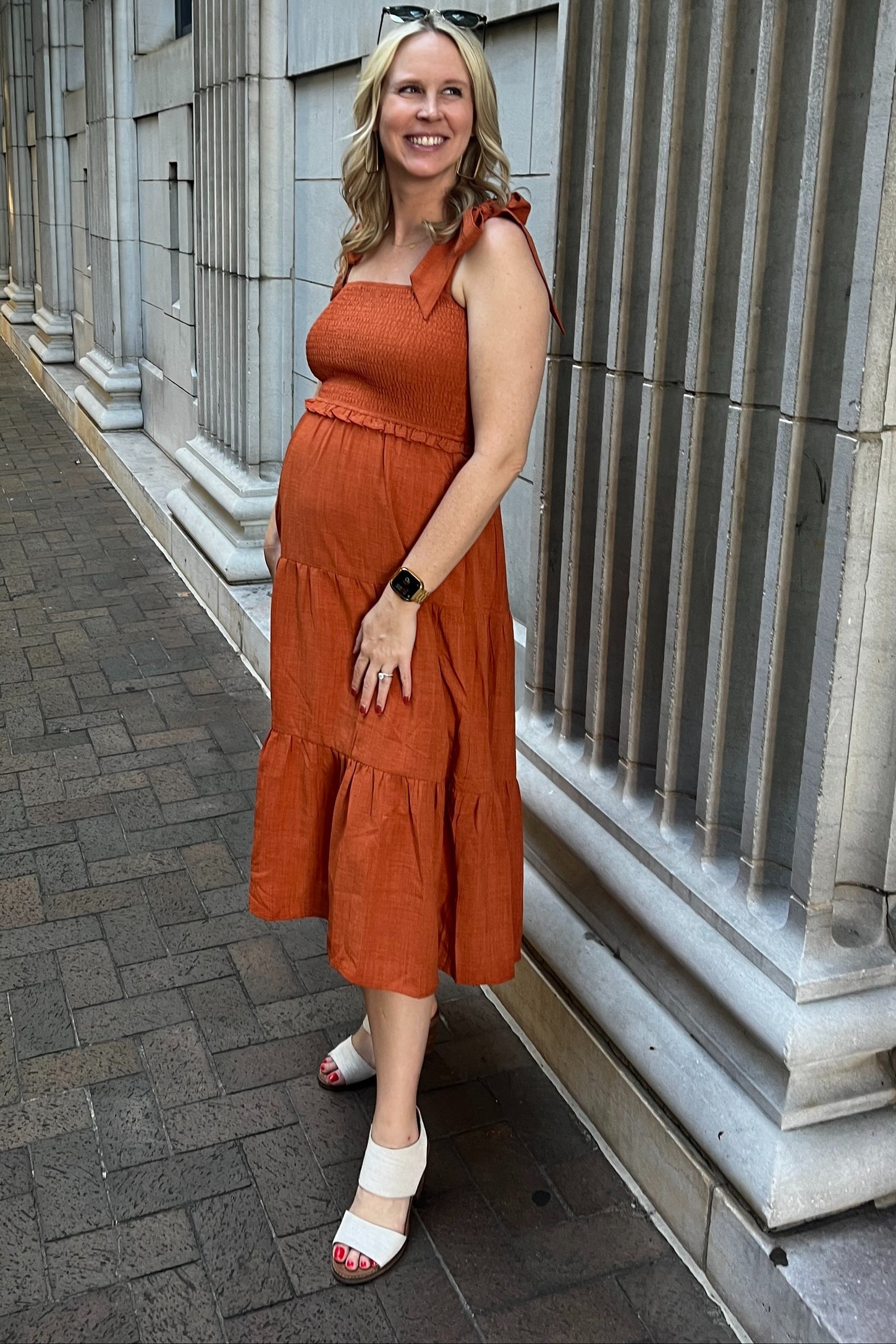 woman wearing an orange maternity sundress with ruffled tiers and a baby bump, standing against a stone wall