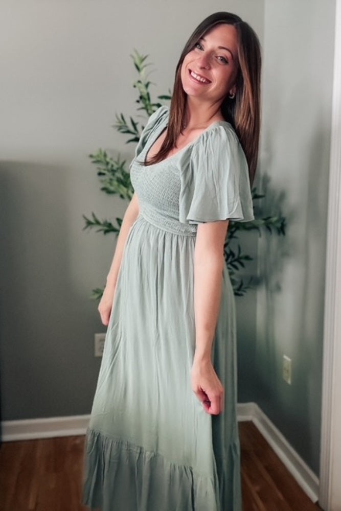 woman wearing a light green maxi dress with short puff sleeves and a smocked bodice, smiling while posing indoors