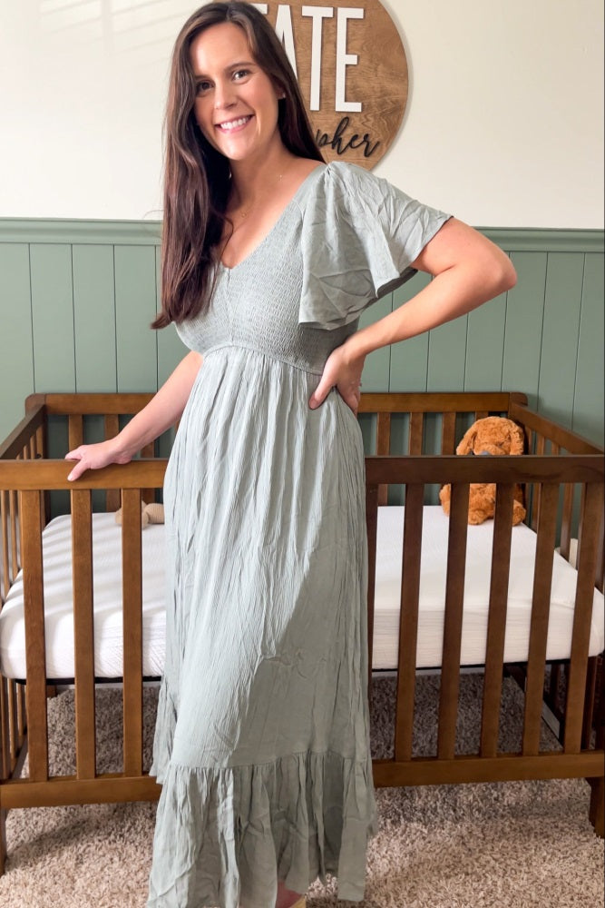 woman wearing a sage green short sleeve maxi dress with a smocked bodice and tiered skirt, standing by a crib