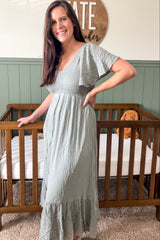 woman wearing a sage green short sleeve maxi dress with a smocked bodice and tiered skirt, standing by a crib