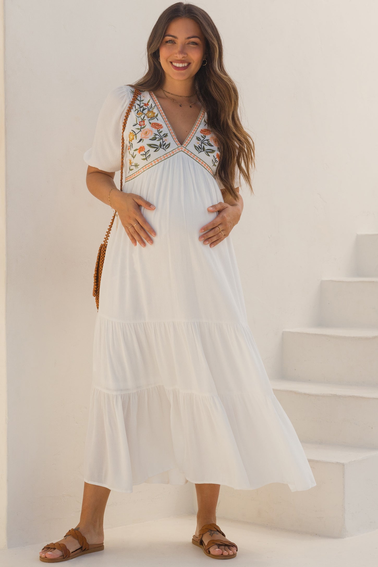 woman wearing a white maternity maxi dress with floral embroidery and a visible baby bump