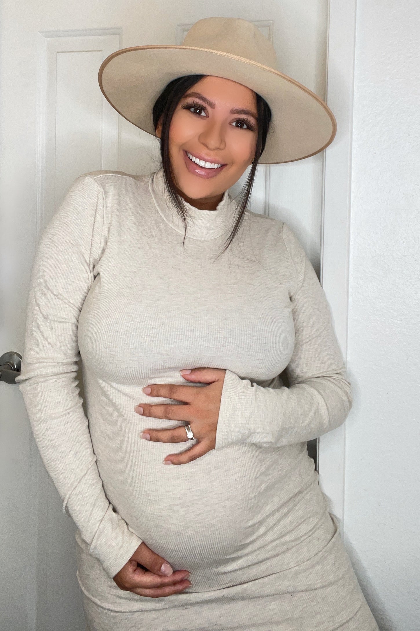 woman wearing a beige ribbed maternity dress and a wide-brimmed hat, holding her baby bump