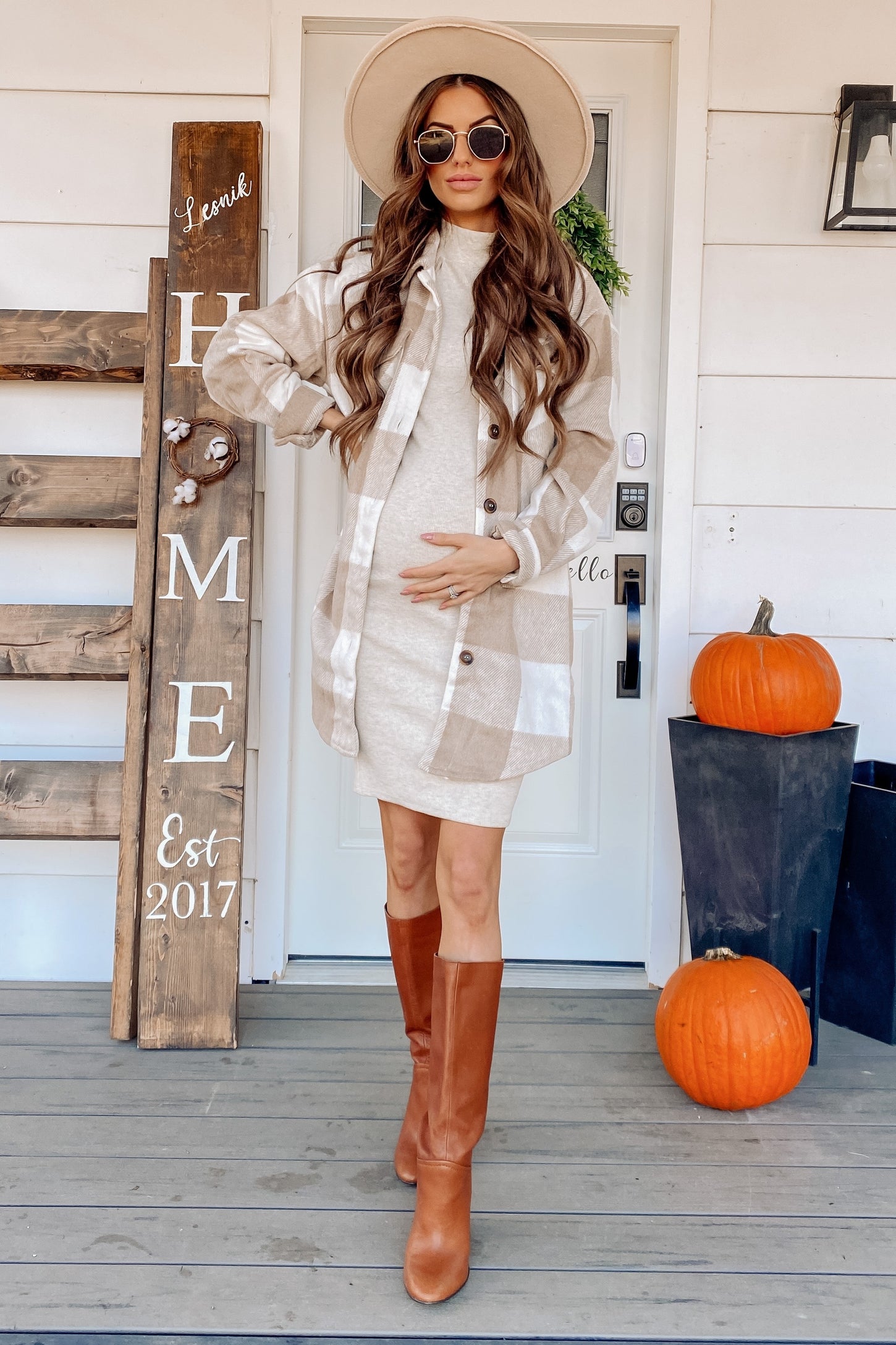 woman wearing a tan and white plaid maternity shacket over a fitted dress, holding her baby bump while standing on a porch