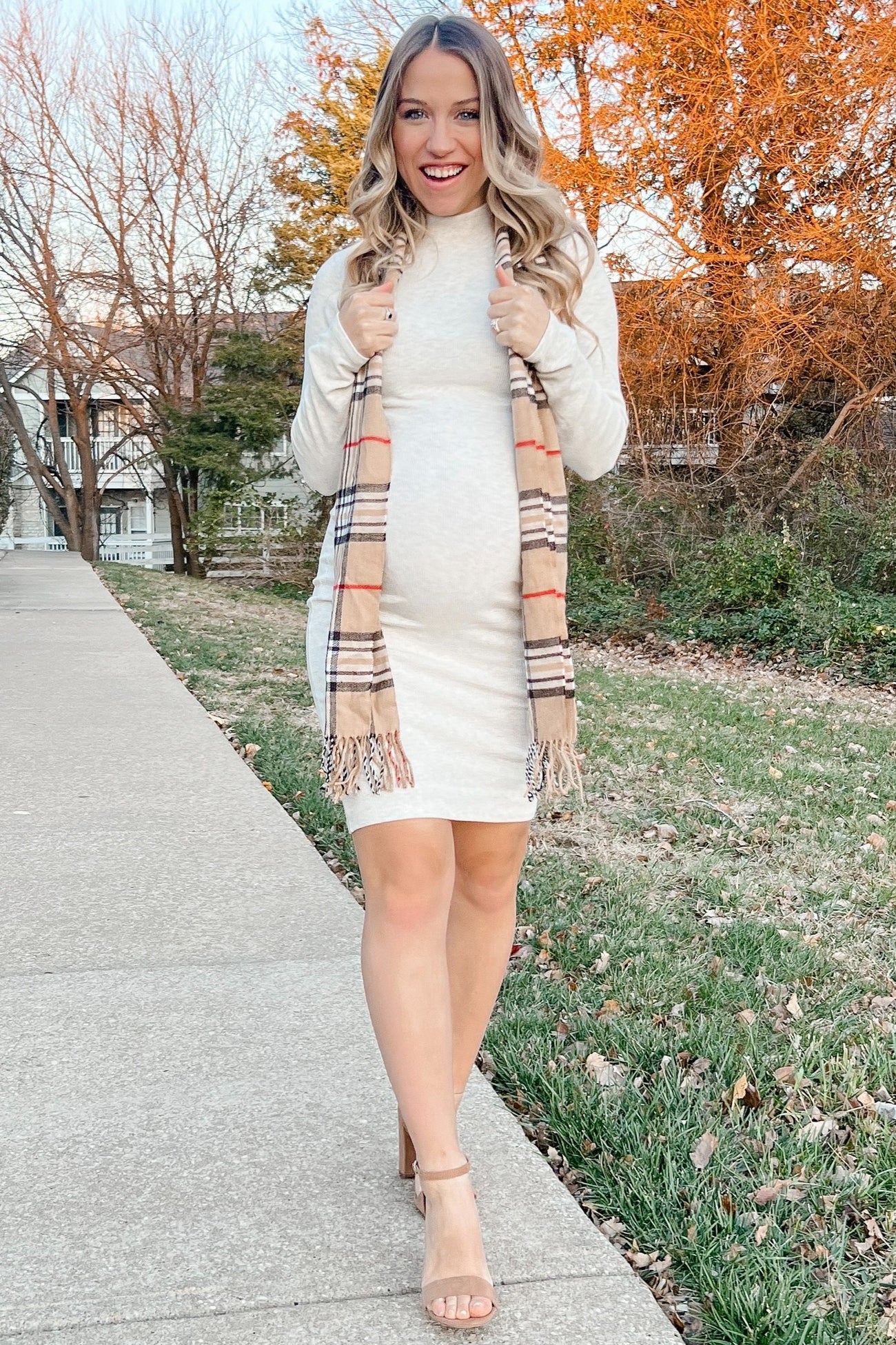 woman wearing a light beige maternity dress with a baby bump and holding a plaid scarf, standing on a sidewalk