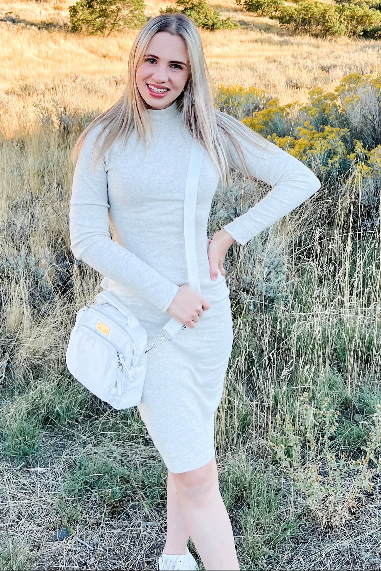woman wearing a light gray long sleeve fitted dress with a drawstring side and a crossbody bag, standing outdoors