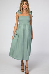 woman wearing a sage green maternity dress with tie shoulder straps and a smocked bodice, featuring a flowing skirt and visible baby bump