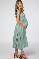 woman wearing a sage green smocked maternity dress with bow tie straps and holding her baby bump