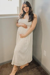 woman wearing a white maternity maxi dress with smocking detail, holding her baby bump while standing by a window