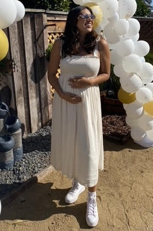 woman wearing a cream maternity dress with smocked bodice and holding her baby bump, styled with white sneakers