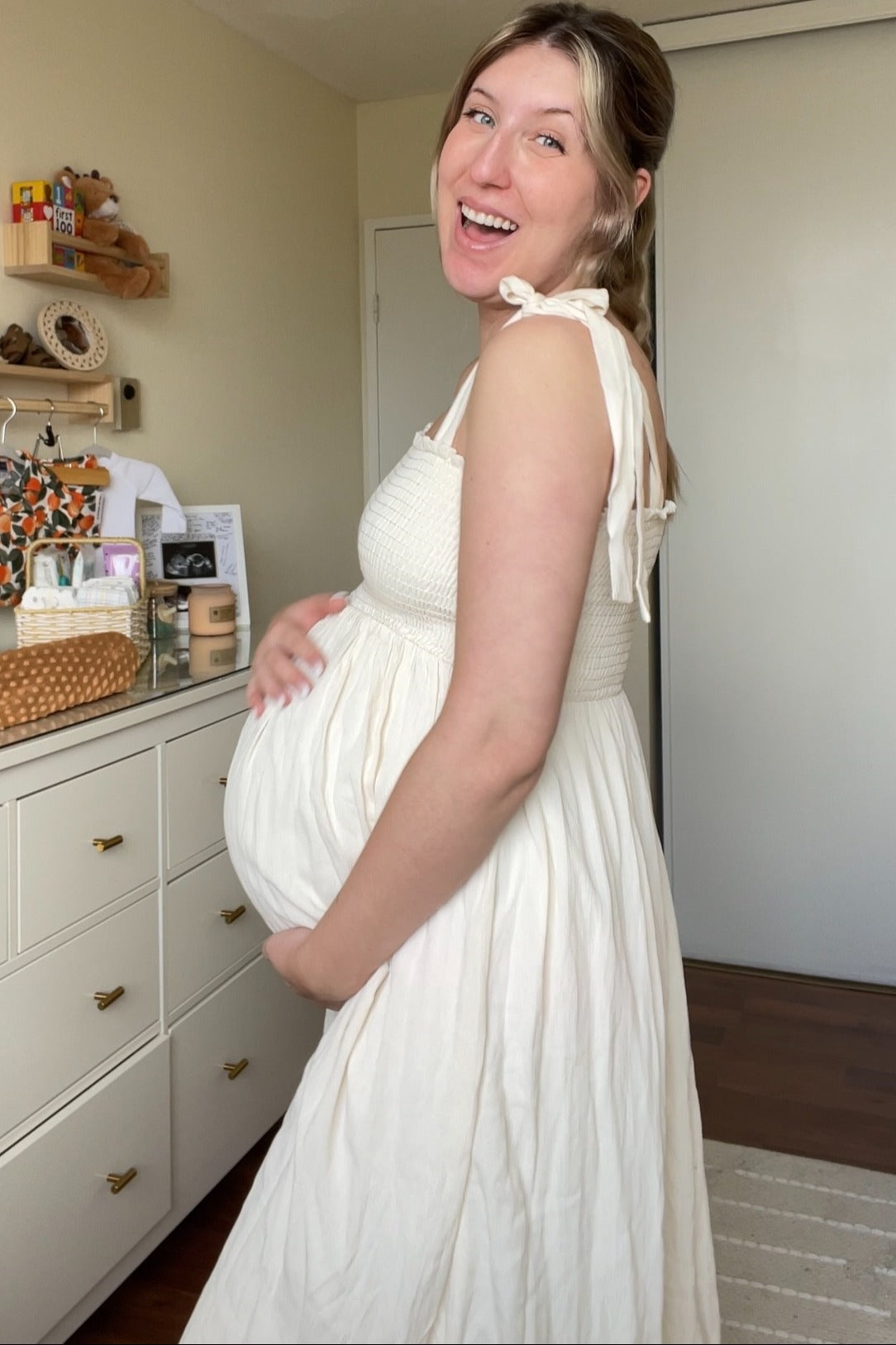 woman wearing a white maternity dress with smocking and tie straps, gently cradling her baby bump