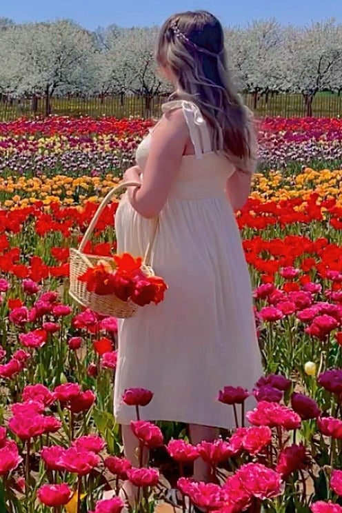woman wearing a cream maternity dress with ruffle straps, holding a basket of flowers and showing her baby bump in a flower field