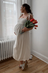 woman wearing a cream maternity maxi dress with smocked bodice, holding a bouquet of tulips and showing her baby bump