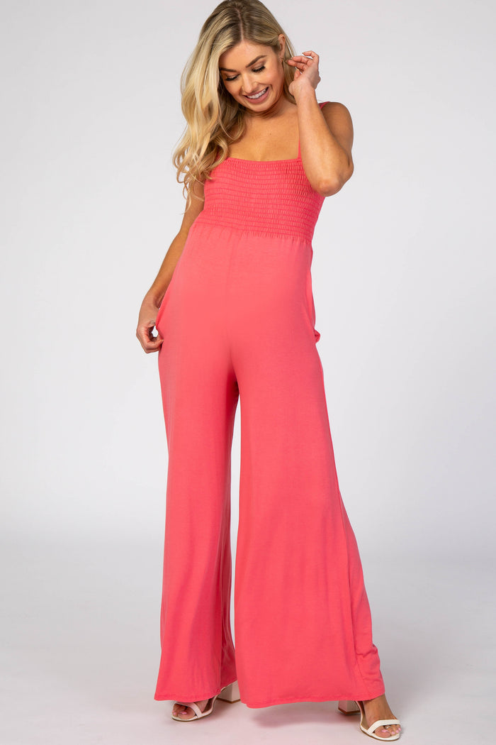 blonde woman wearing a coral pink smocked wide leg maternity jumpsuit with pockets