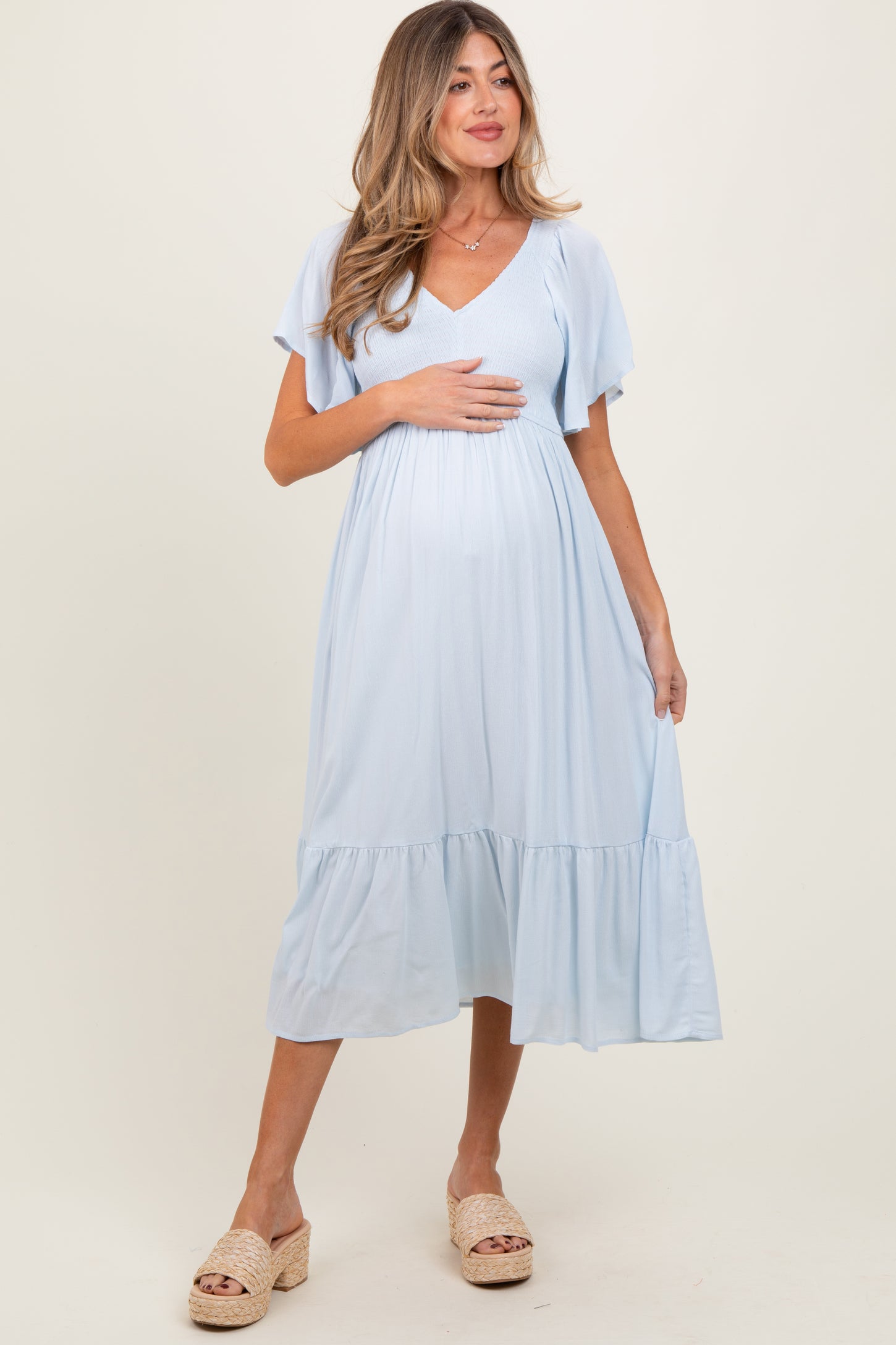 woman wearing a light blue maternity dress with short flutter sleeves and a tiered hem, cradling her baby bump