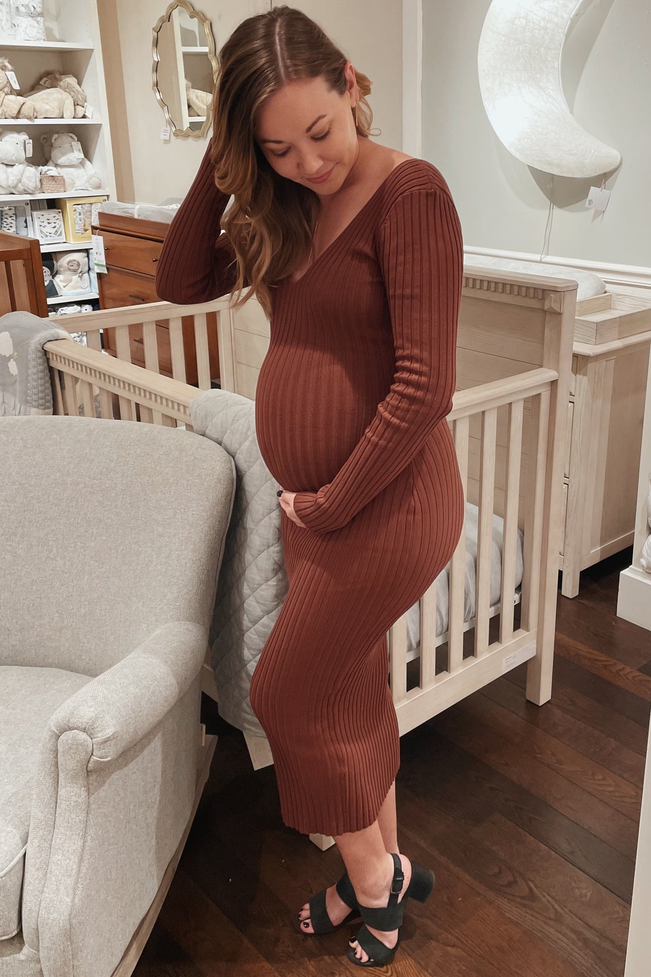 woman wearing a rust ribbed maternity dress and cradling her baby bump while standing near a crib