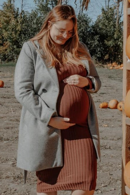 pregnant woman in a brown dress and grey coat holding her belly at a pumpkin patch