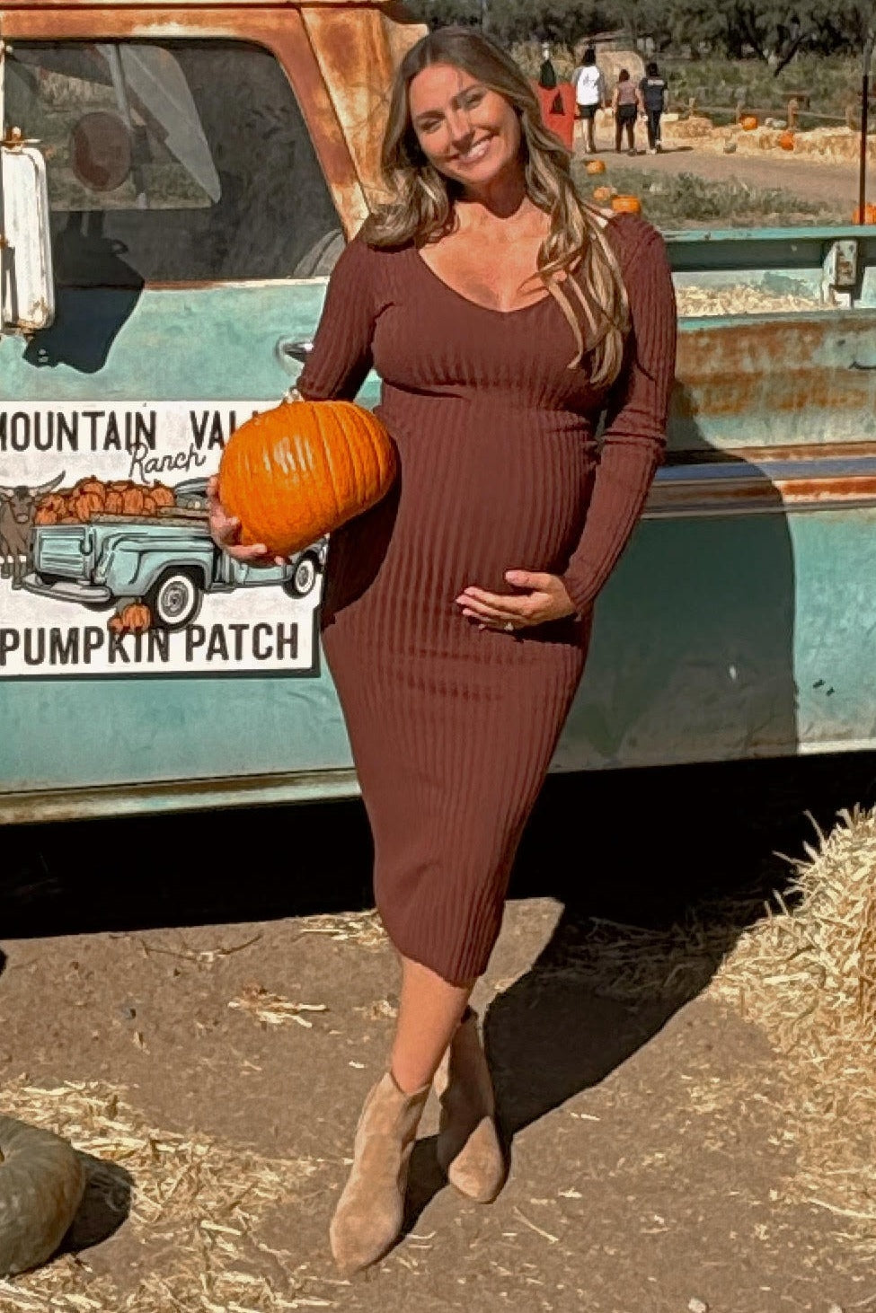 Smiling pregnant woman in a brown ribbed midi dress holding a pumpkin at a pumpkin patch by a vintage truck.