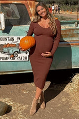 Smiling pregnant woman in a brown ribbed midi dress holding a pumpkin at a pumpkin patch by a vintage truck.