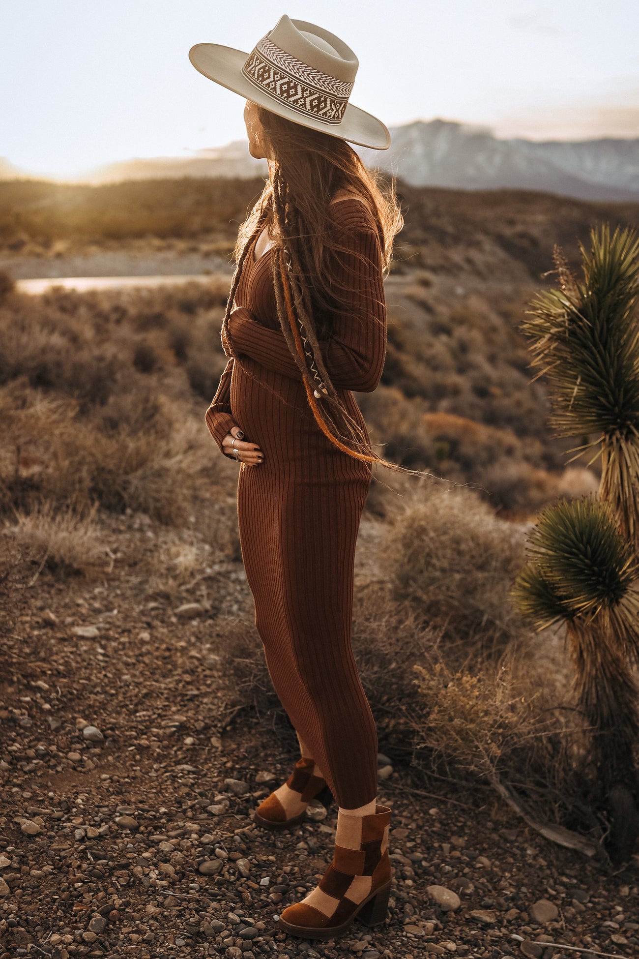 woman wearing a rust ribbed maternity set and holding her baby bump, accessorized with a wide-brimmed hat