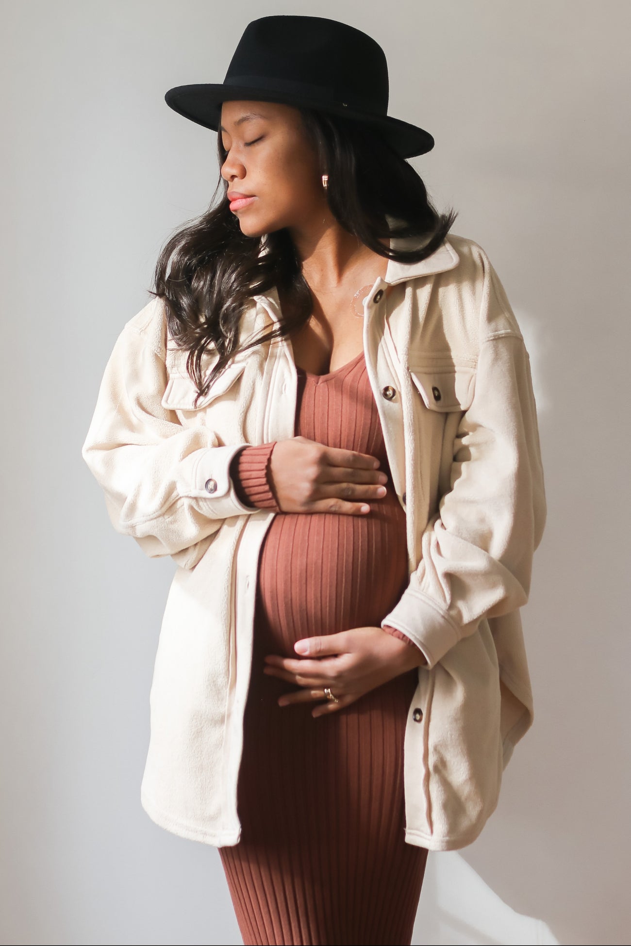 Pregnant woman in a black hat and cream shacket holding her baby bump in a ribbed maternity dress
