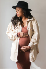 Pregnant woman in a black hat and cream shacket holding her baby bump in a ribbed maternity dress