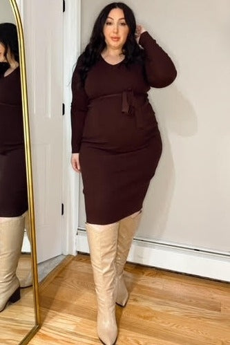 woman wearing a dark brown maternity dress with a tie detail and tall beige boots, showing baby bump in front of a mirror