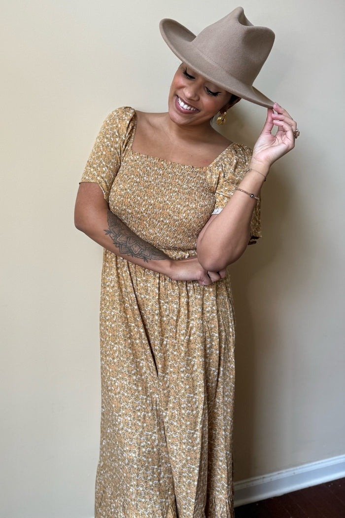 woman wearing a mustard floral maxi dress with short sleeves and a square neckline, smiling and holding a beige hat