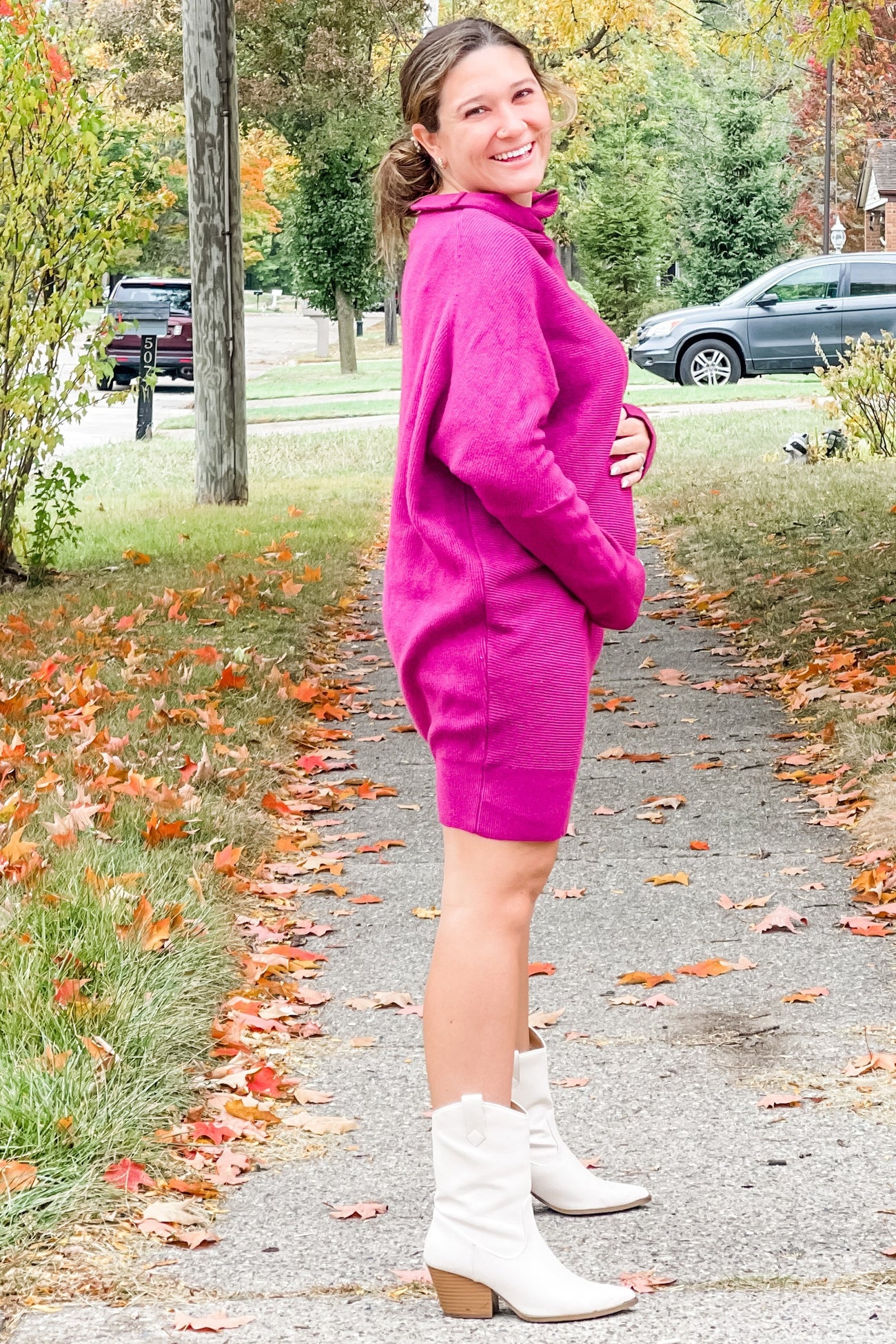 woman wearing a fuchsia maternity sweater dress and holding her baby bump, standing on a leaf-covered path