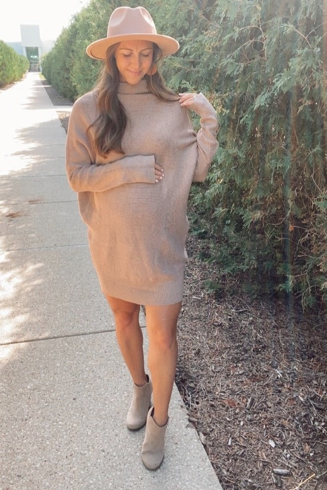 woman wearing a taupe oversized maternity sweater dress and a beige hat, gently holding her baby bump outdoors