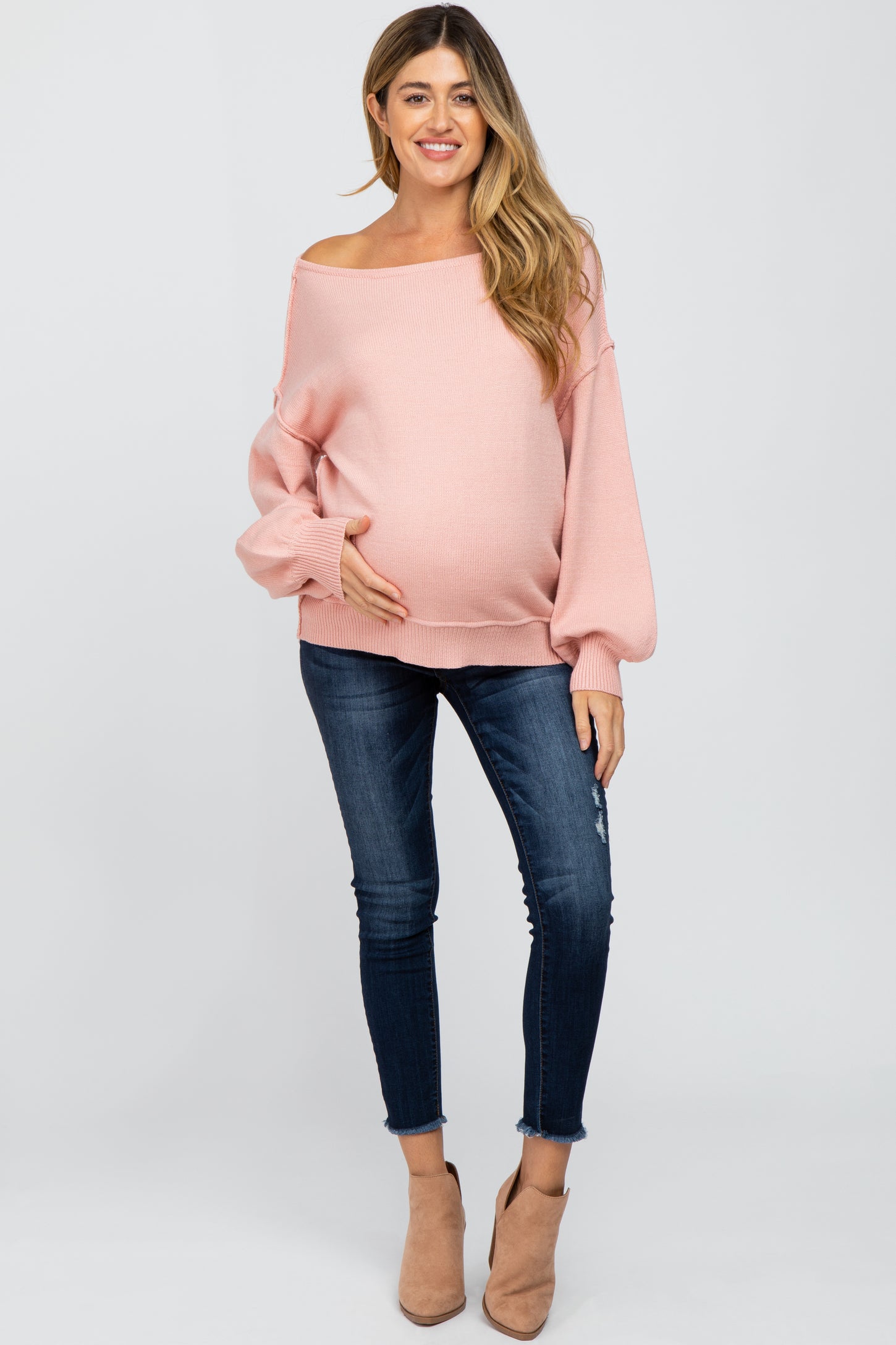 woman wearing a pink off-the-shoulder maternity sweater and holding her baby bump with fitted jeans and ankle boots