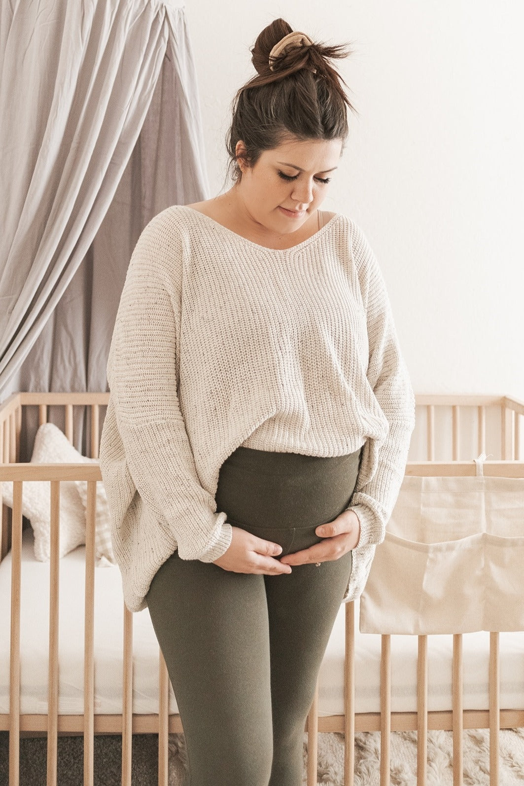 pregnant woman in cream sweater and olive leggings holding her belly while standing in front of a wooden crib