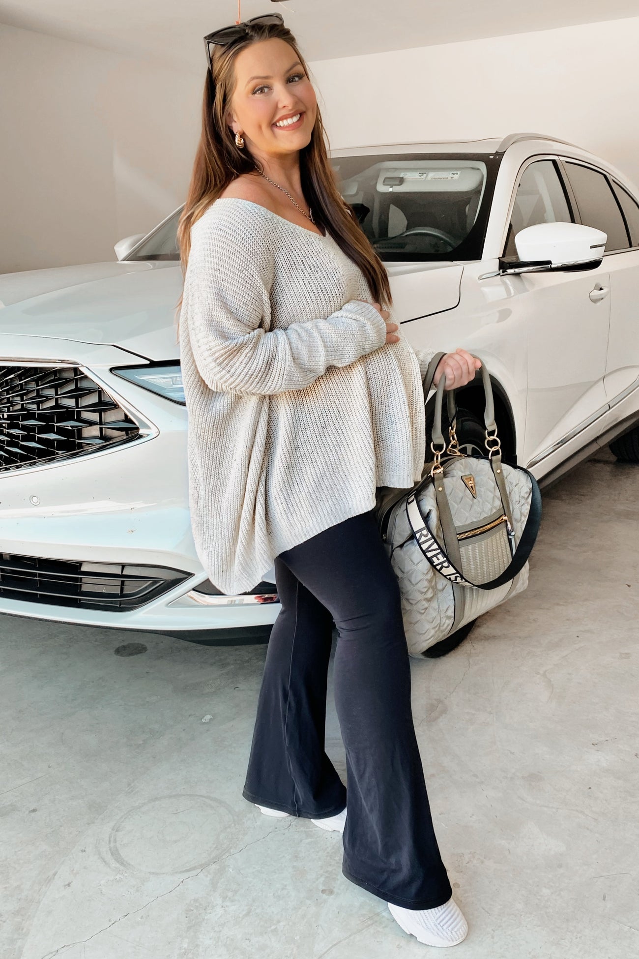 woman wearing a light gray oversized sweater and black maternity pants, holding a handbag and showing her baby bump