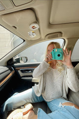 woman wearing a light beige maternity sweater and sitting in a car, holding a teal camera, with ripped light blue jeans on