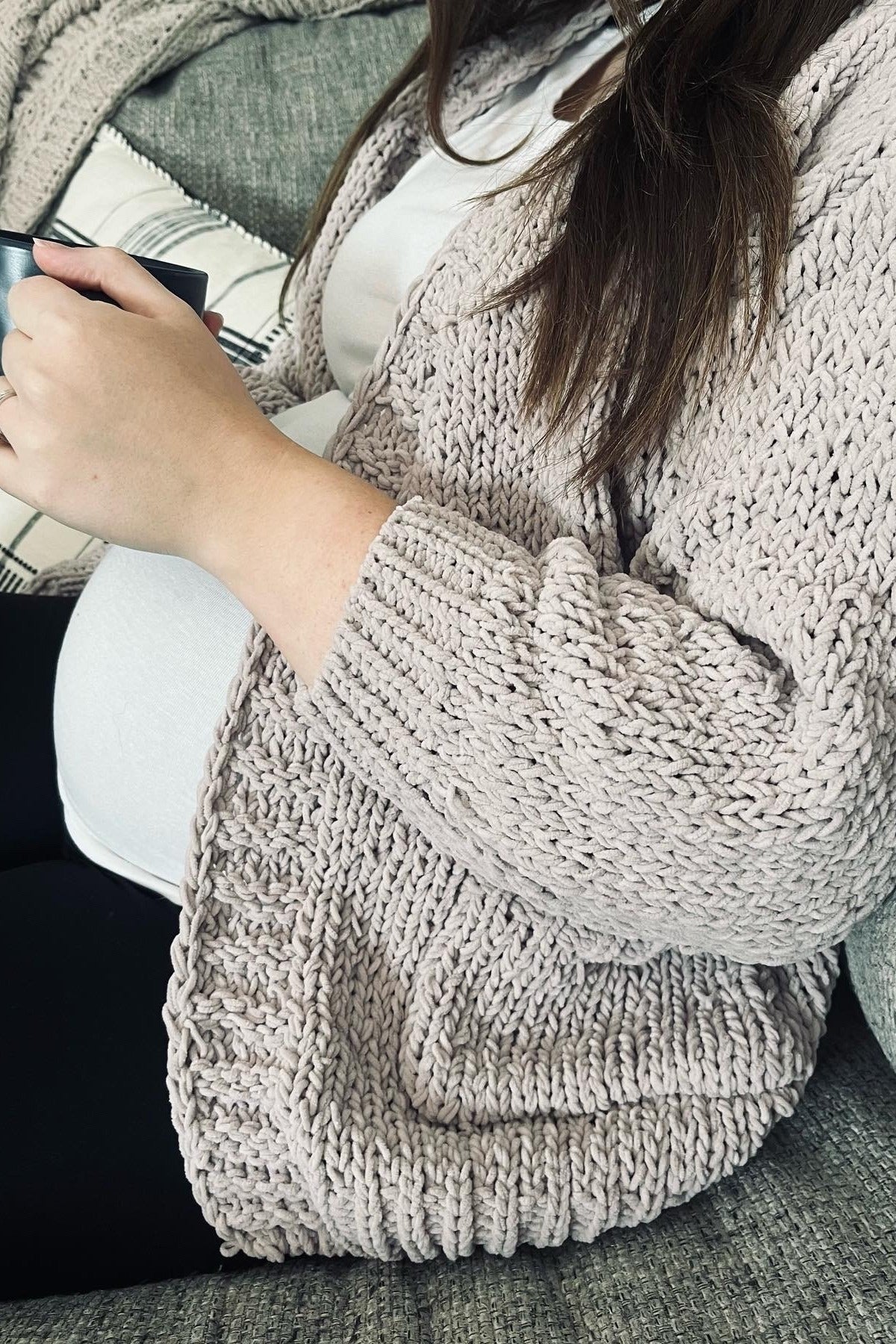 woman wearing a soft pink knitted maternity cardigan and black leggings, holding a mug, with a visible baby bump