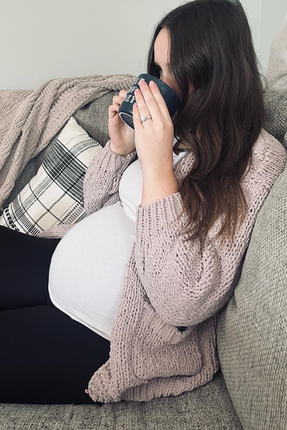 woman wearing a light pink knitted cardigan and a white maternity top, sitting on a couch with a baby bump visible and holding a coffee mug