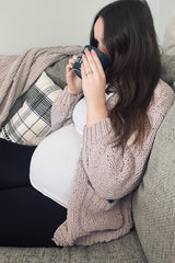 woman wearing a light pink knitted cardigan and a white maternity top, sitting on a couch with a baby bump visible and holding a coffee mug