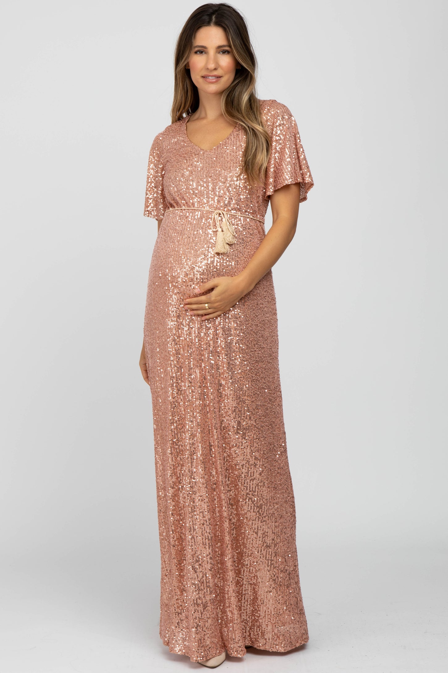 woman wearing a rose gold sequin maternity gown with a v-neck and belted waist, holding her baby bump