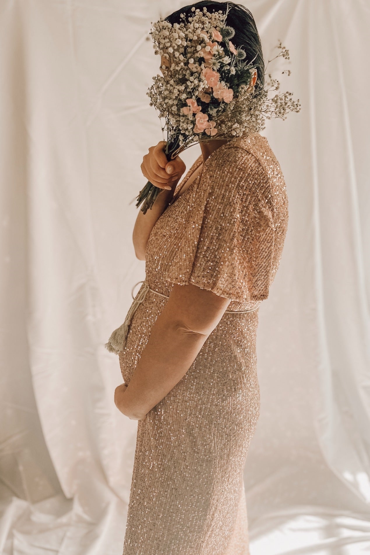 woman wearing a rose gold sequined maternity dress, holding a bouquet of flowers and showing her baby bump